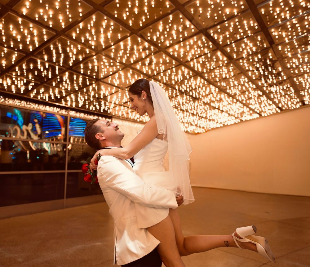 Groom lifts bride in wedding dress under sparkling ceiling lights in romantic venue.