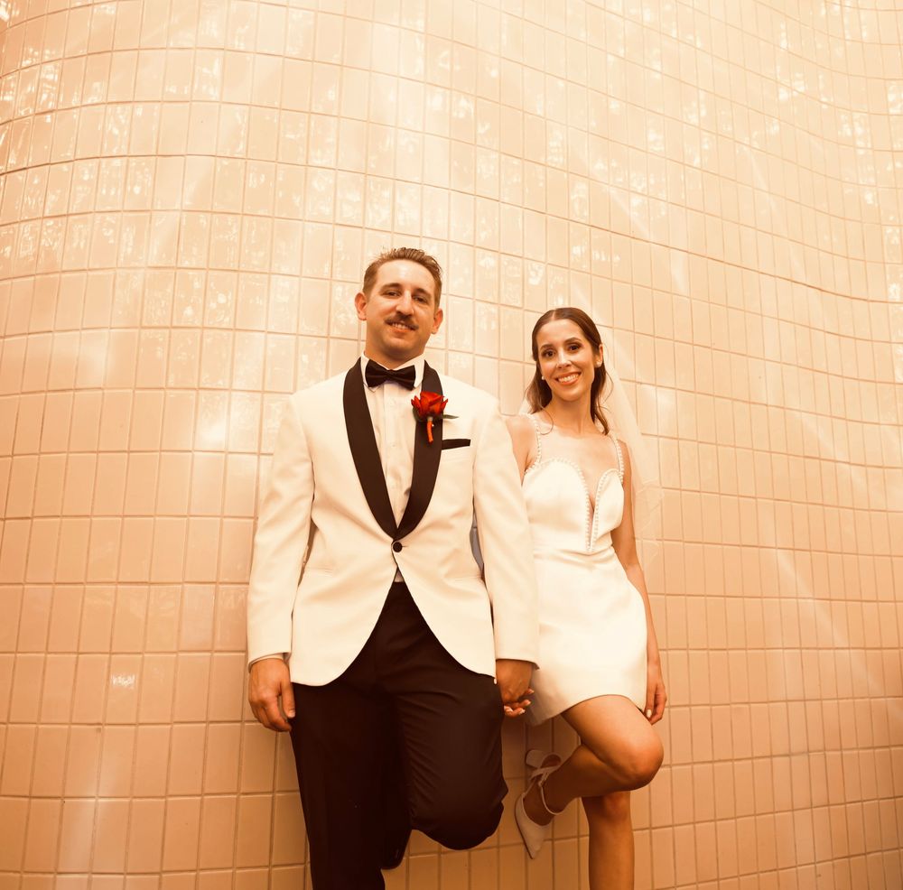 Couple in wedding attire posing against a pink tiled wall, groom in white tuxedo and bride in short white dress.