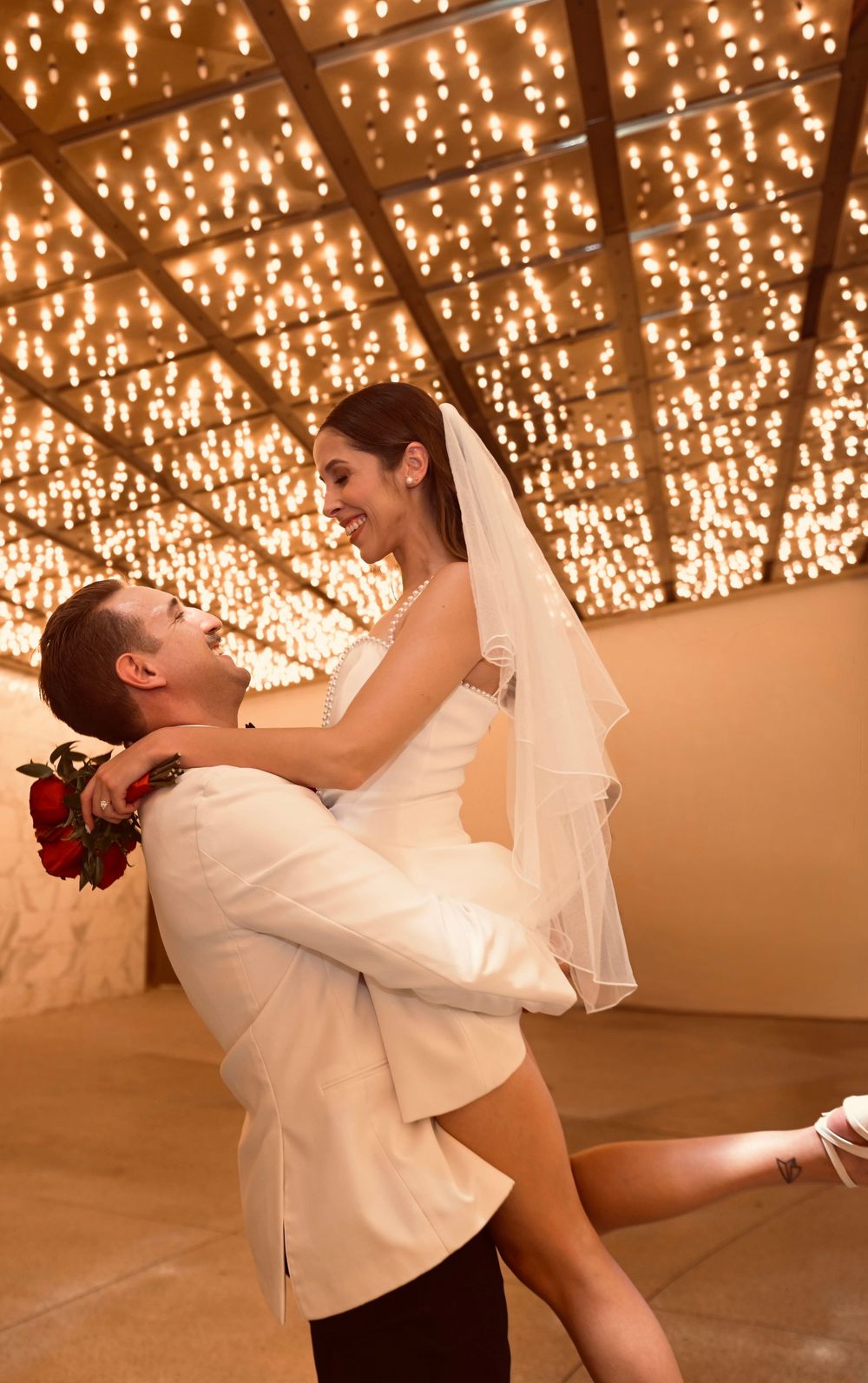 Groom lifting bride in white dress and veil under sparkling lights at wedding reception.