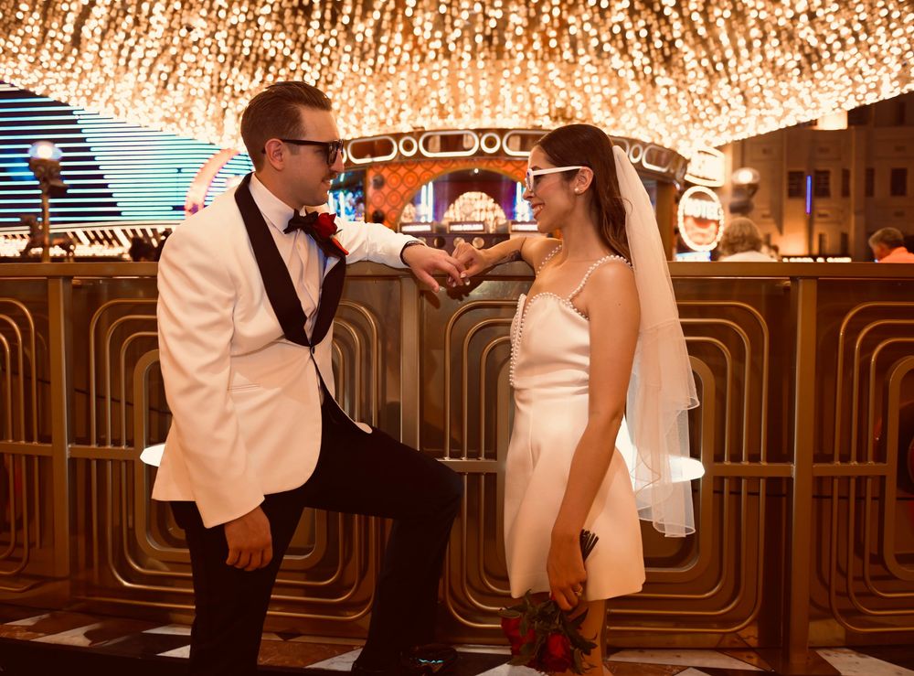 Bride and groom in white attire making heart shape with hands under casino lights in Las Vegas.
