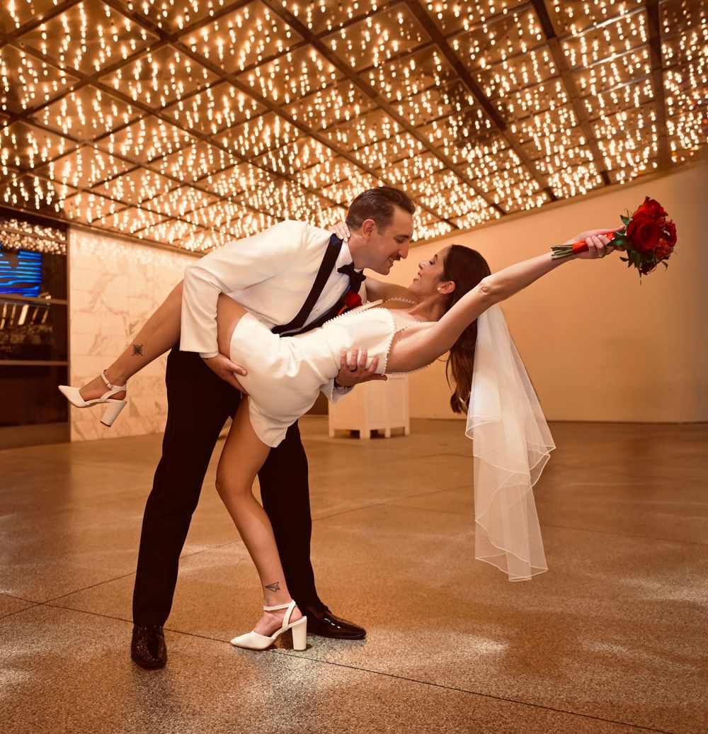 Bride and groom share a romantic dip kiss on a dance floor under a canopy of warm twinkling lights.