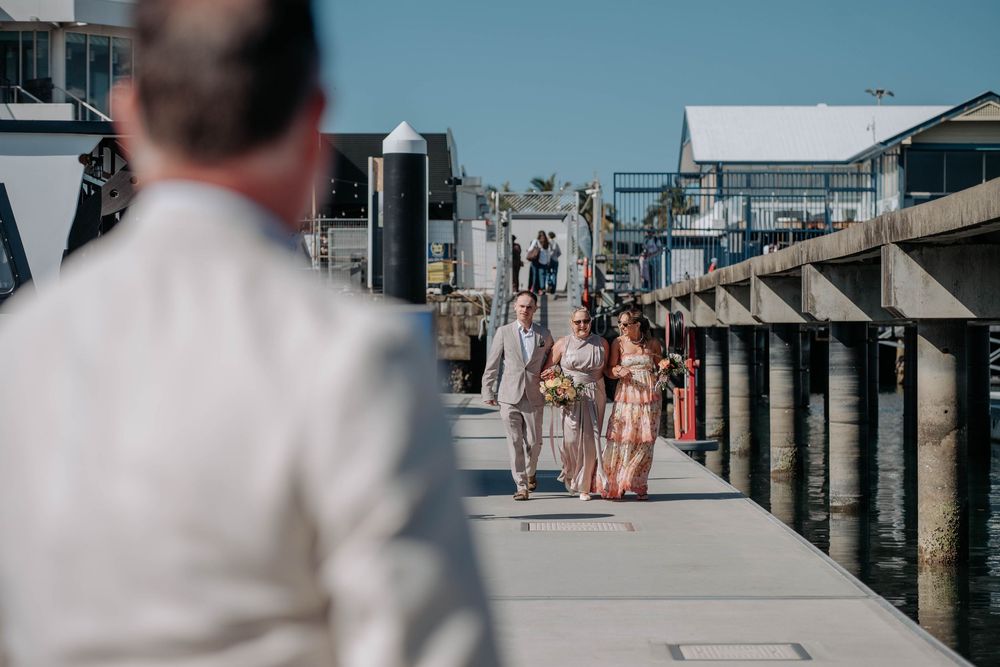 Bridal party walking down a sunny marina dock toward a groom in a white suit at a waterfront wedding.