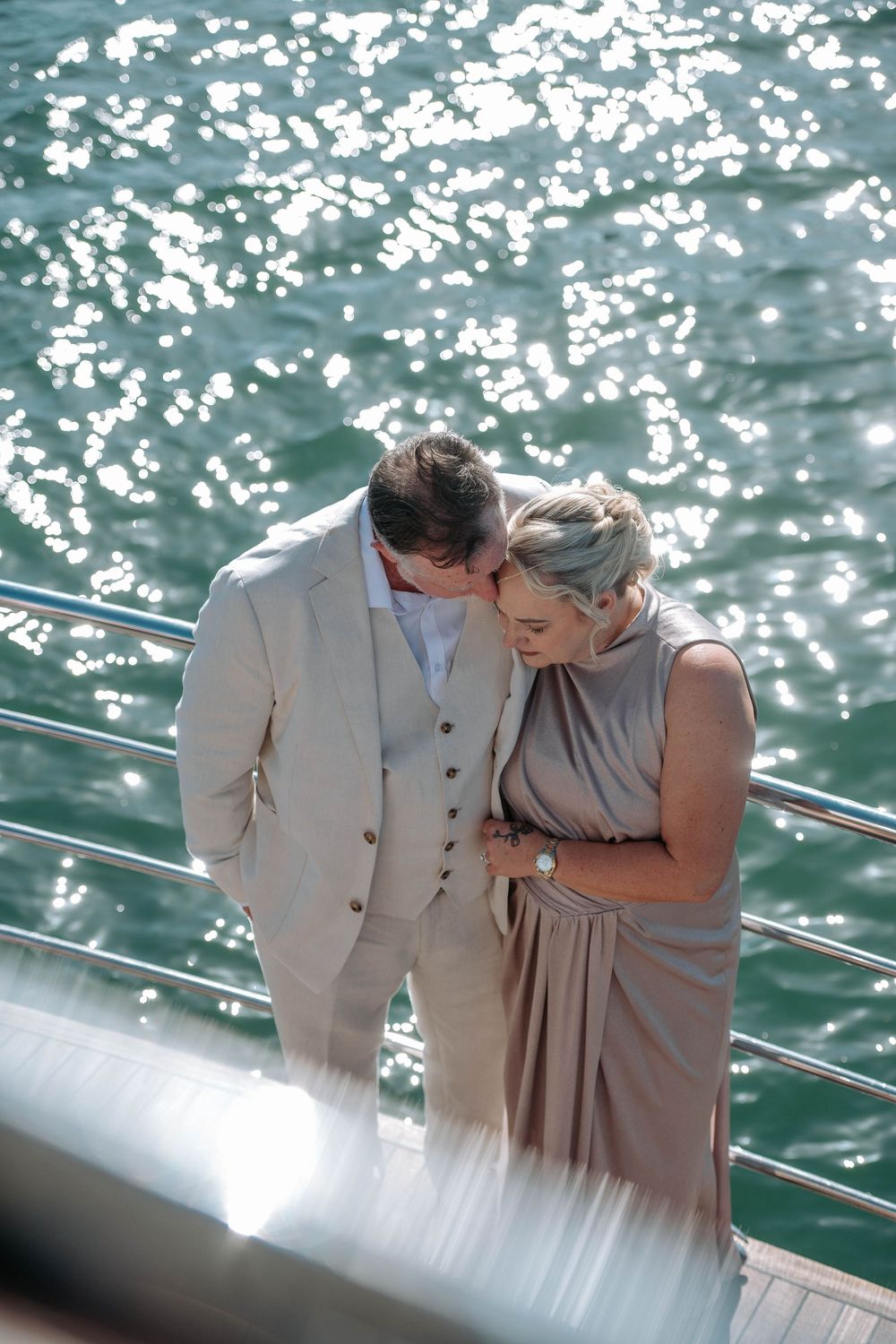 Couple in elegant beige attire sharing a tender moment on a yacht deck with sparkling water behind them.