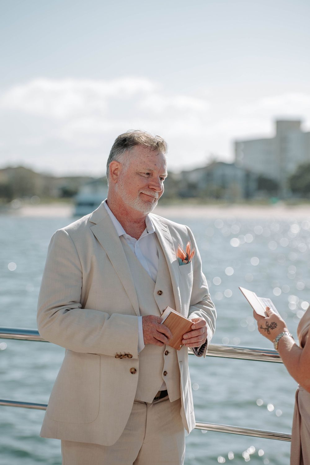 Groom in beige suit smiling during outdoor waterfront wedding ceremony on a sunny day.