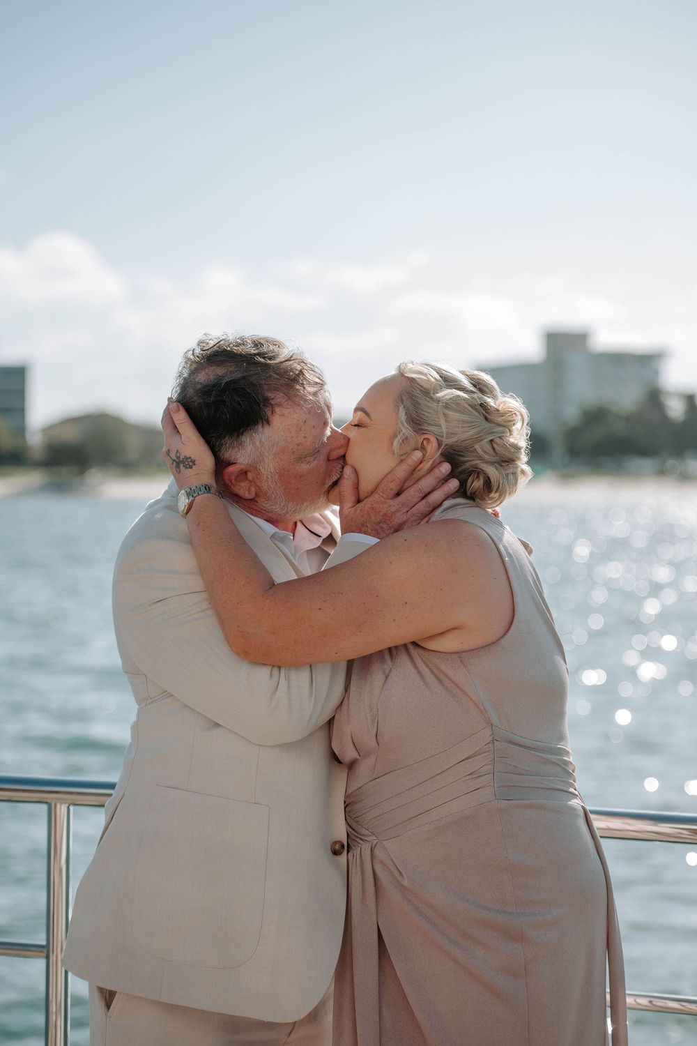 Elderly couple sharing a romantic kiss on a yacht deck with sparkling ocean and city skyline in the background.