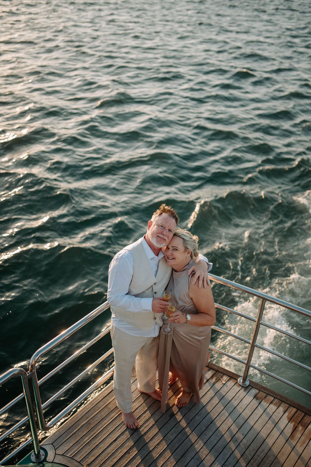 Couple embracing on yacht deck at sunset with ocean waves in background, wearing elegant beige and white outfits.