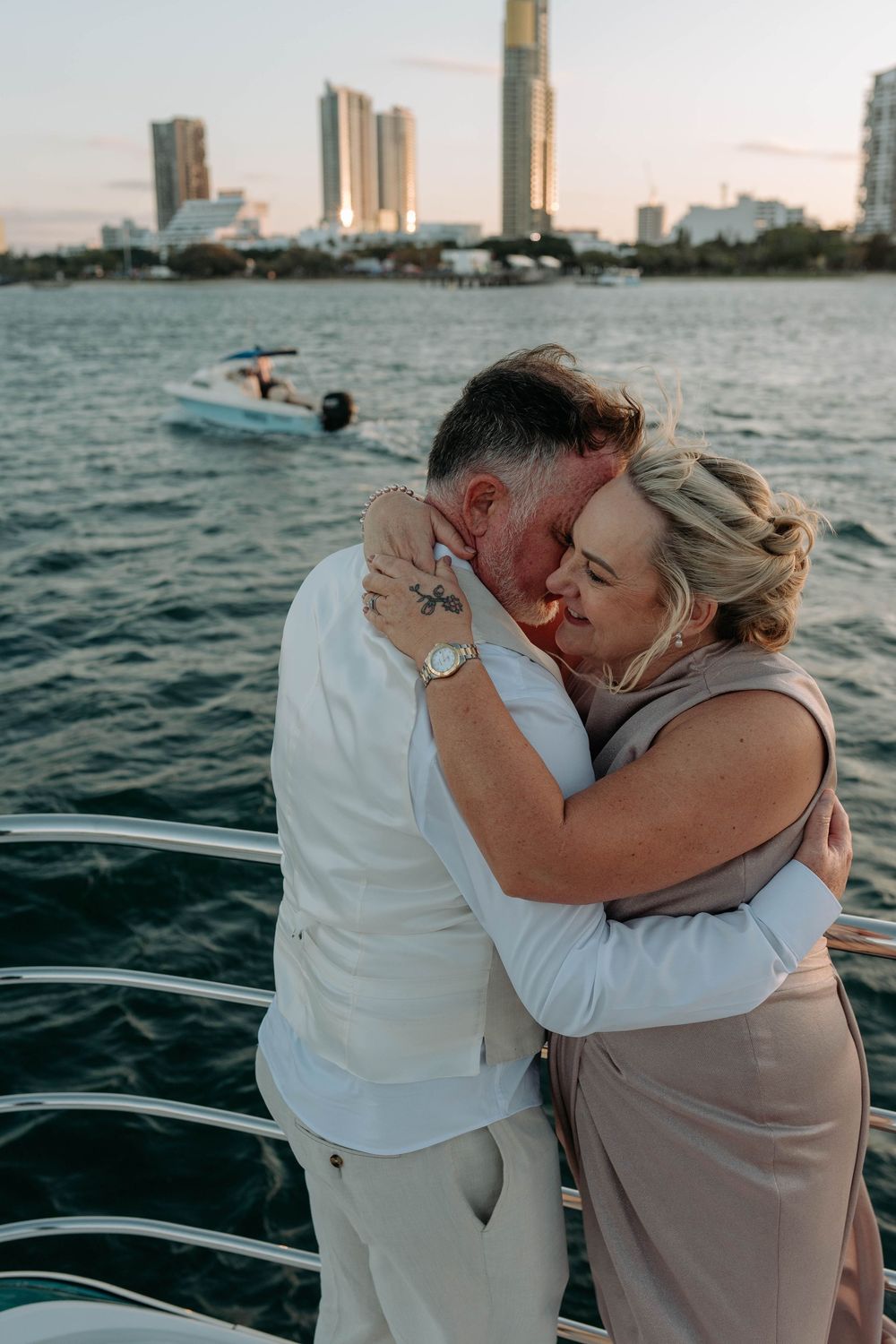 Couple embracing and kissing on a yacht at sunset with city skyline and water in the background.