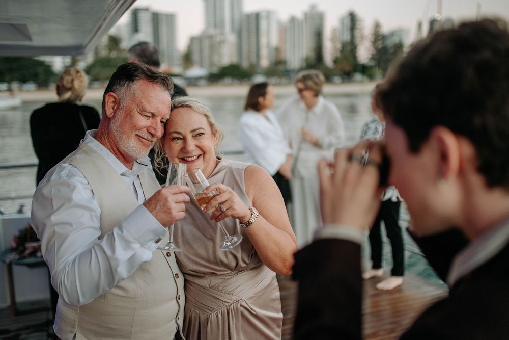 Couple laughing and toasting drinks at an outdoor waterfront wedding reception while a photographer captures the moment.