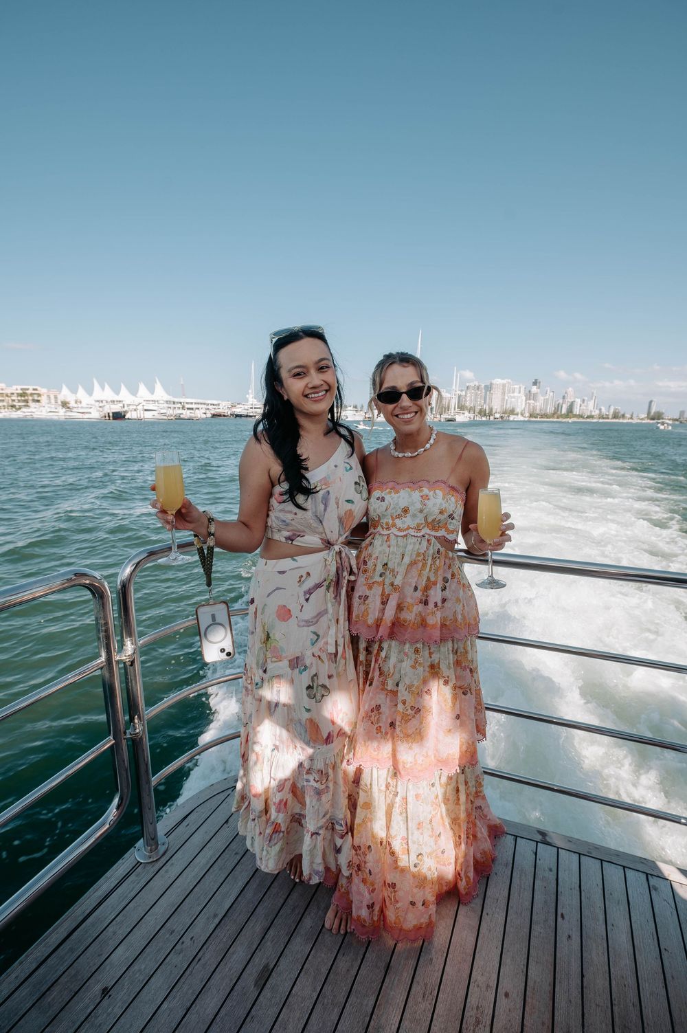 Two women in floral dresses holding champagne glasses on a yacht deck with city skyline in background.