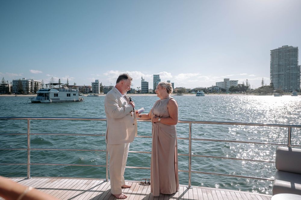 Couple exchanging vows on a luxury yacht deck with sparkling waterway and city skyline backdrop on a sunny day.