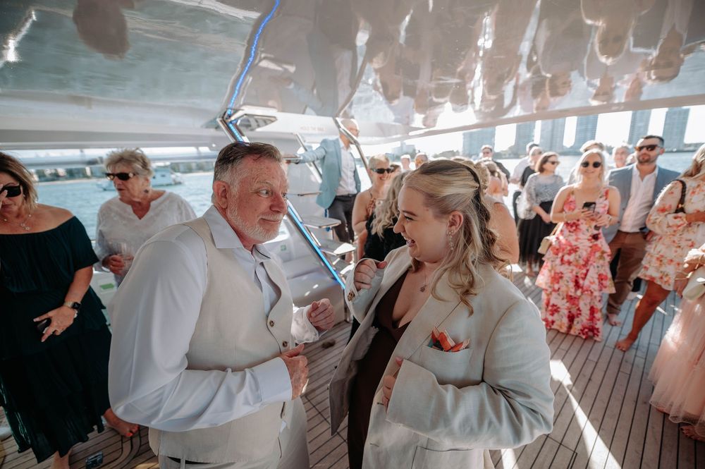 Couple in white attire smiling at each other during a wedding celebration on a sunny boat deck.
