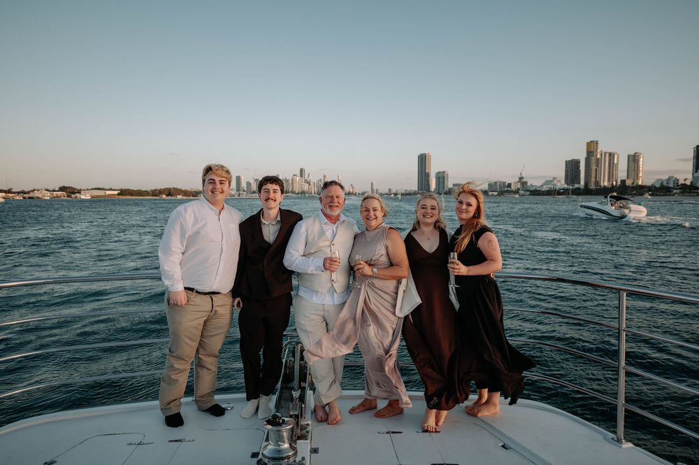 Six people posing together on a yacht with a city skyline and water in the background at sunset.