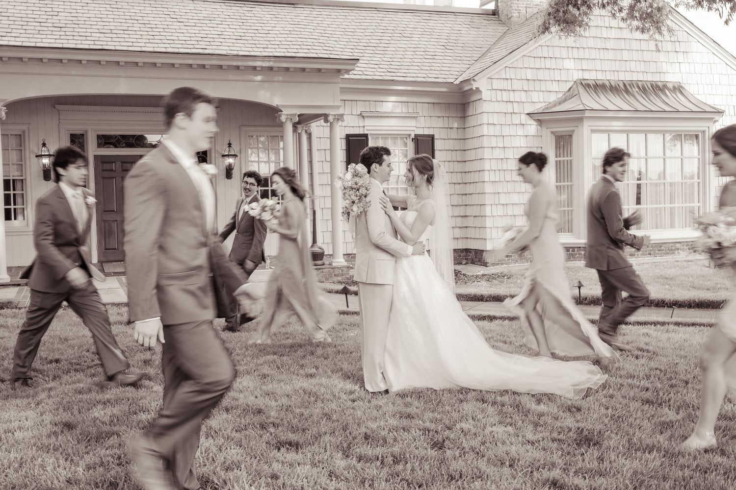 Black and white photo of a wedding party walking across a lawn in front of a charming house.