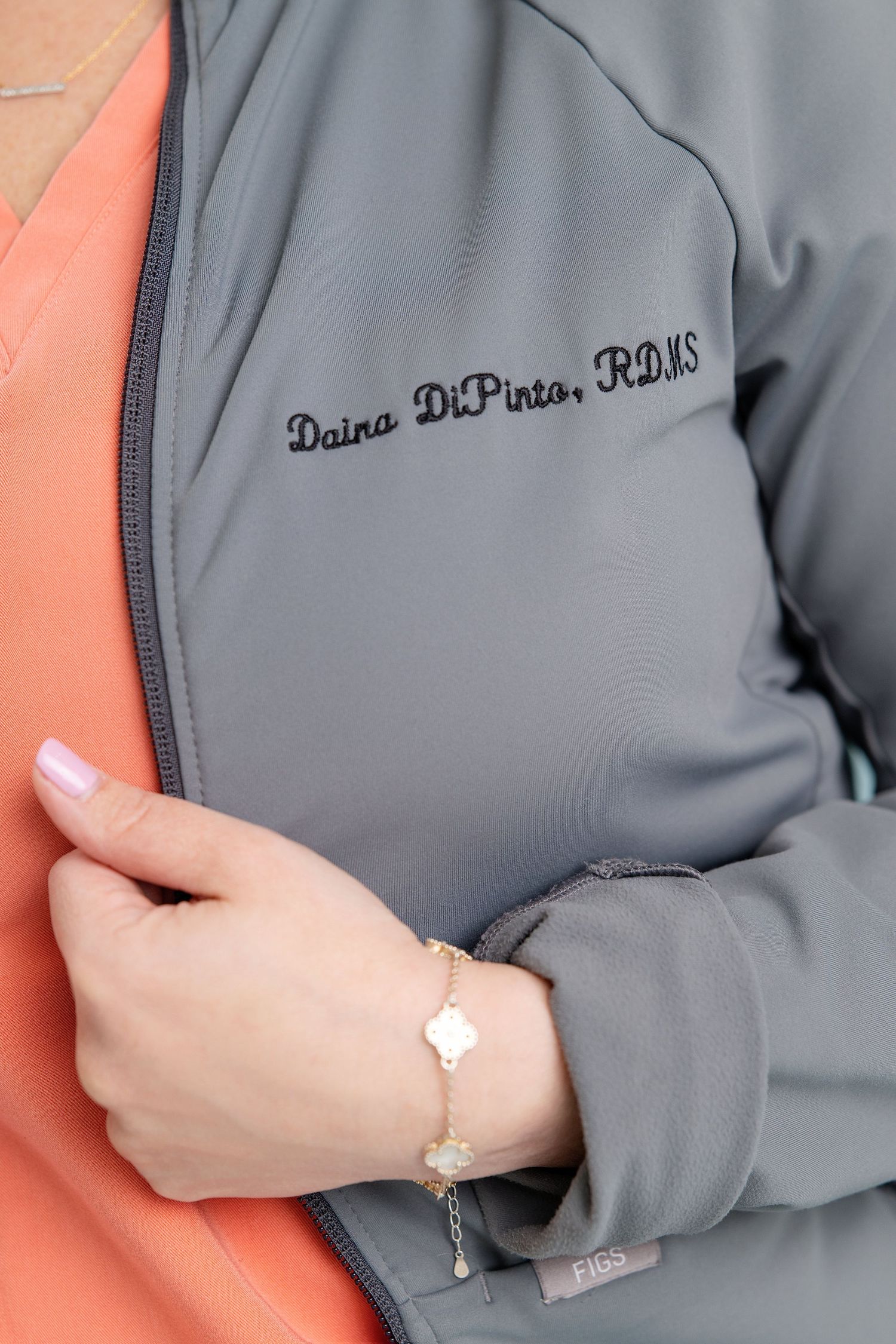 Gray zip-up jacket embroidered with Daina DiPinto, RDMS, worn with a coral top and delicate gold bracelet.