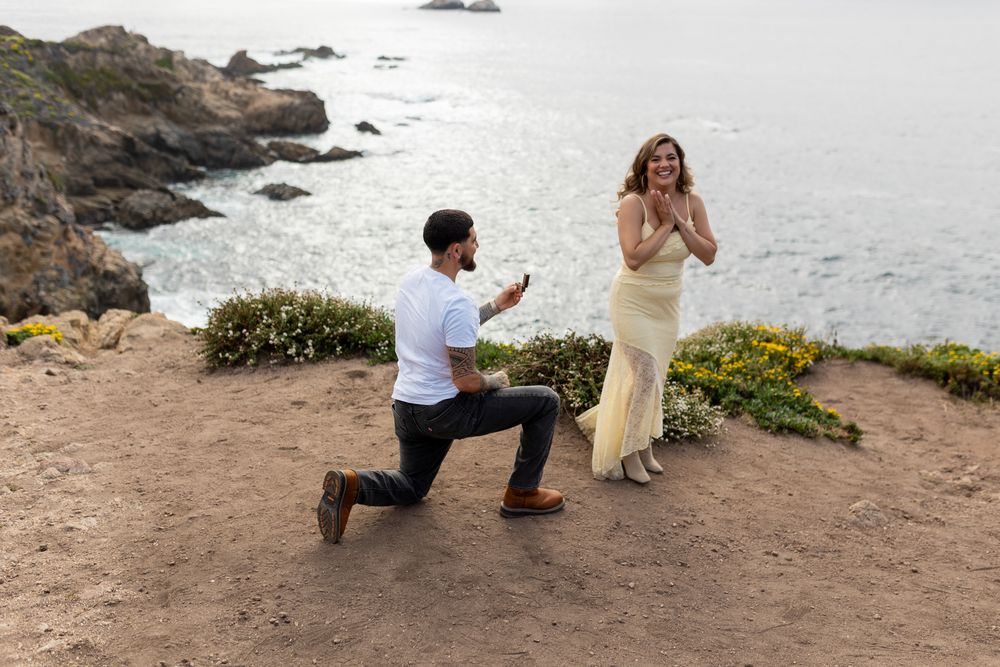 Man proposing on one knee to woman in yellow dress on coastal cliff with ocean views.