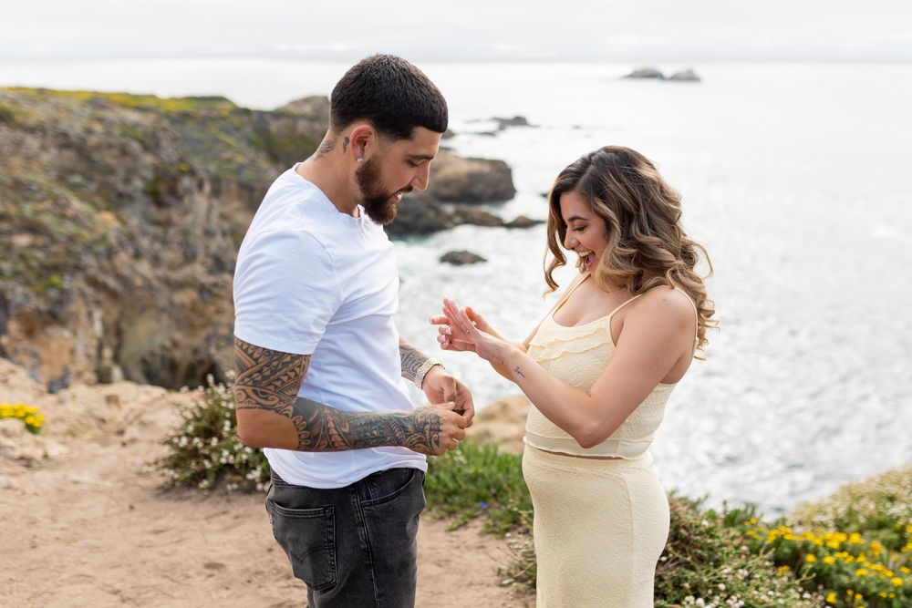 A tattooed man places a ring on a woman's finger during a cliffside proposal with ocean views.