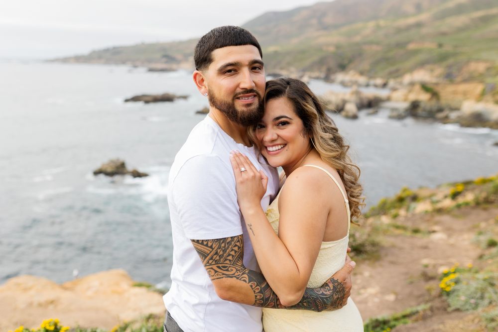 Couple embracing on a coastal cliff with ocean views, man with tattoos in white tee hugging smiling woman in yellow dress.