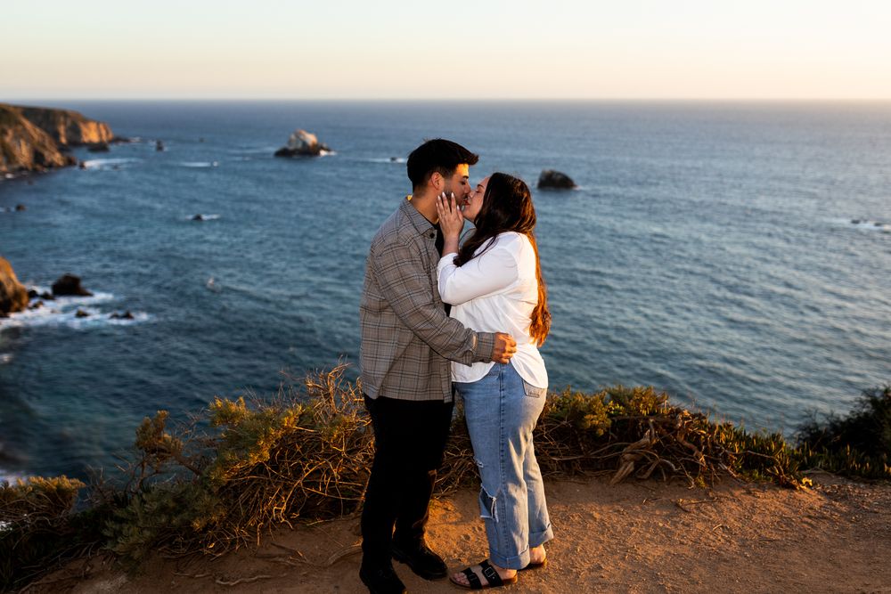 Couple embracing and kissing on a coastal cliff at sunset with blue ocean and rocky shoreline in the background.
