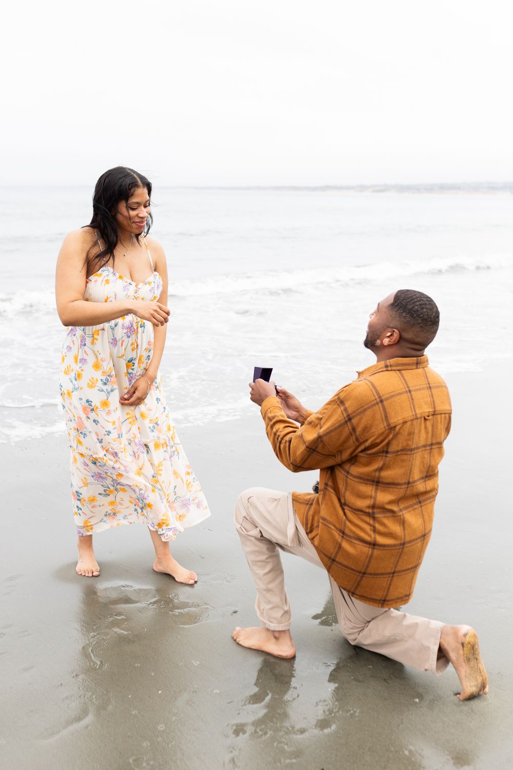 Man proposing on one knee to woman in floral dress on a beach, holding open ring box by ocean waves.
