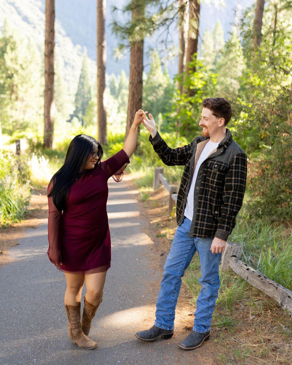 Couple dancing on a forest path surrounded by tall pine trees and mountains, woman in burgundy dress and man in plaid shirt.