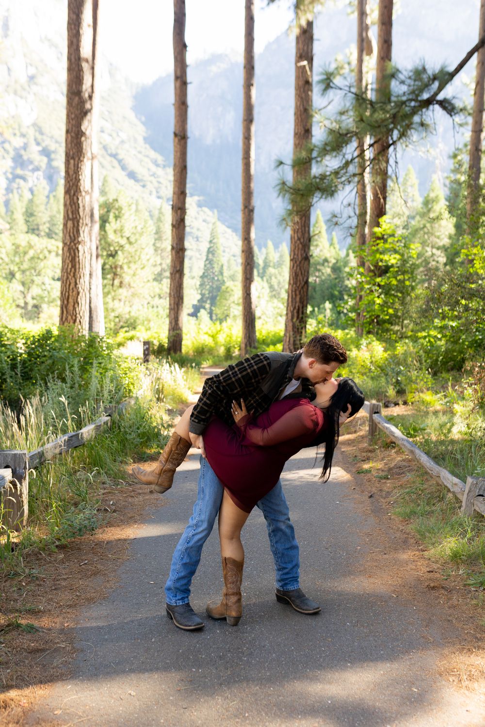 Couple sharing a romantic dip kiss on a forest path surrounded by tall pines and mountain views.