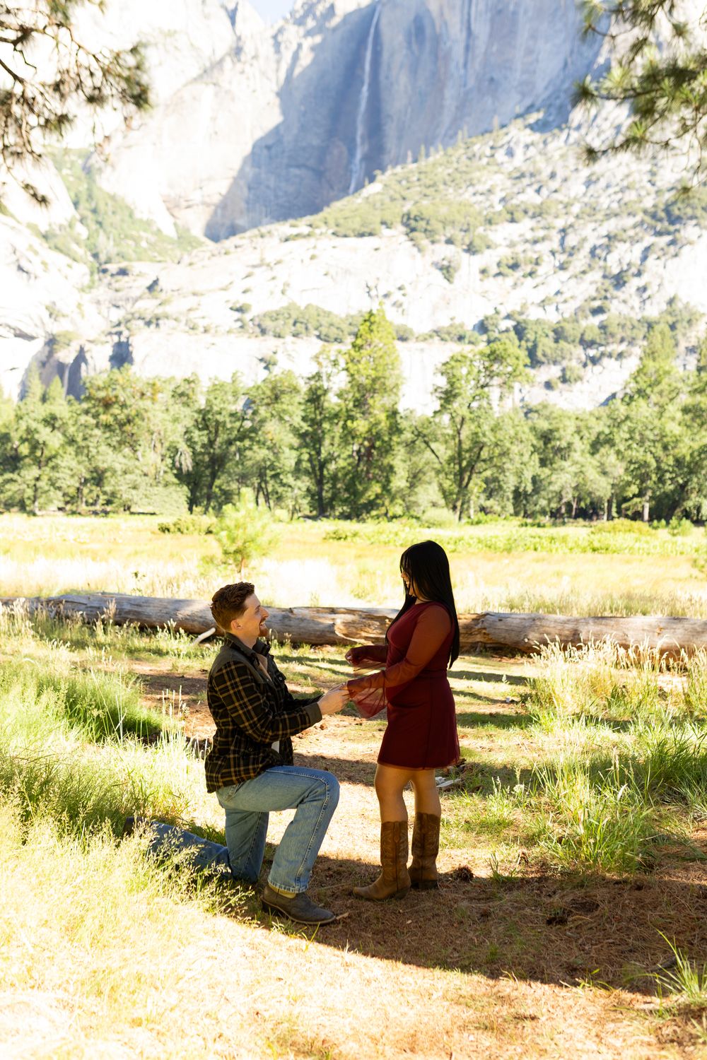 Man kneeling on one knee proposing to woman in red dress at Yosemite National Park with waterfall backdrop.