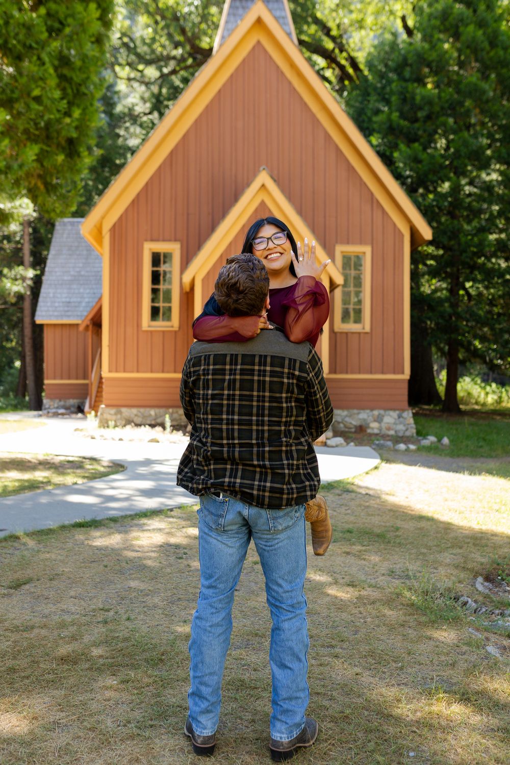 Couple embracing in front of a charming wooden chapel surrounded by trees, woman smiling and waving.