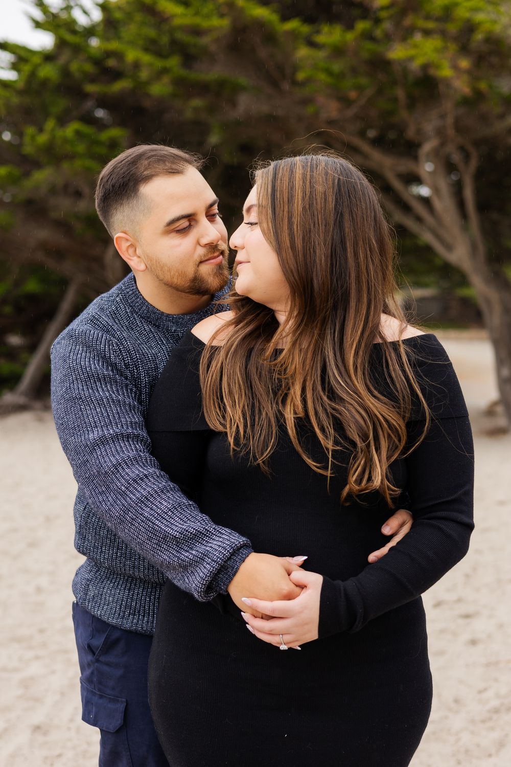 A couple shares a tender moment on a sandy beach, the man in a blue sweater embracing a woman in a black dress.