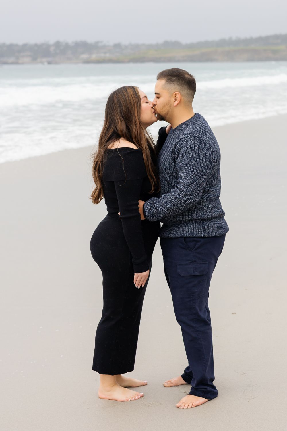Couple sharing a romantic kiss on a sandy beach, woman in black outfit and man in grey sweater and navy pants.