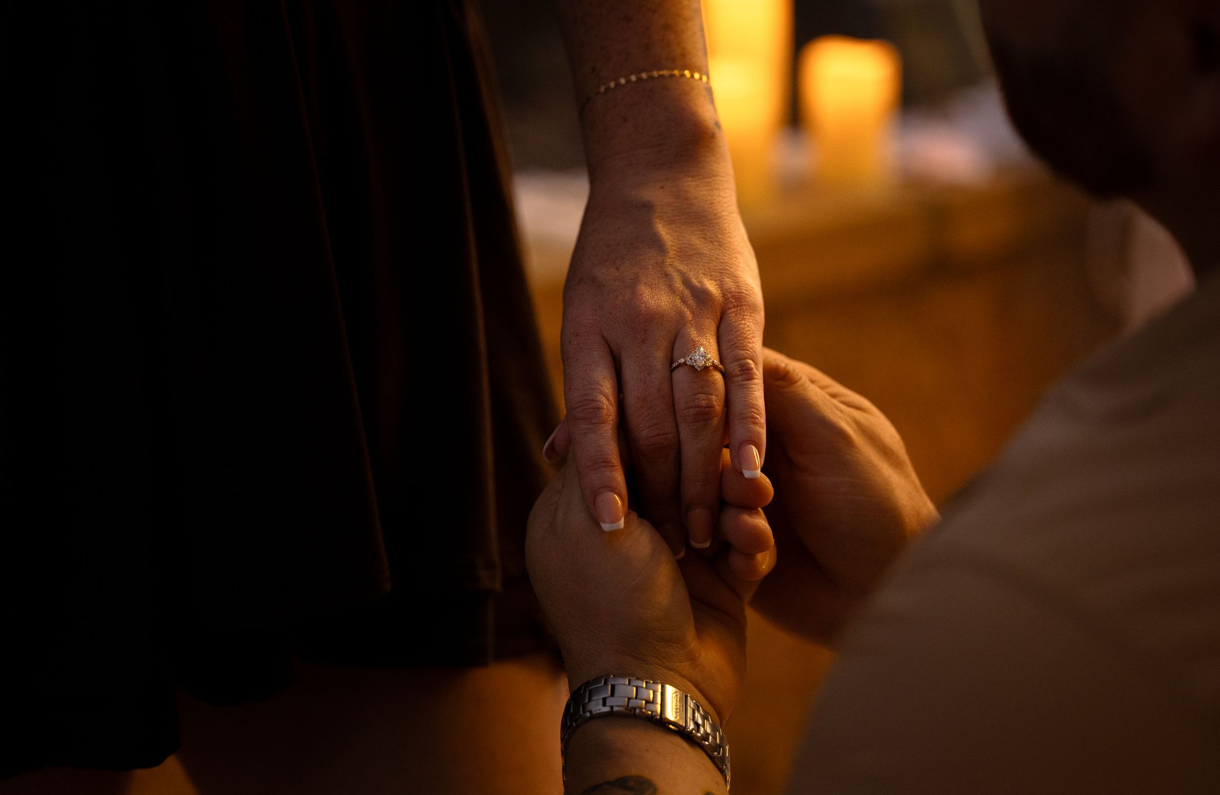 Two people holding hands by candlelight, one wearing a ring and bracelet, in a romantic warm-toned setting.