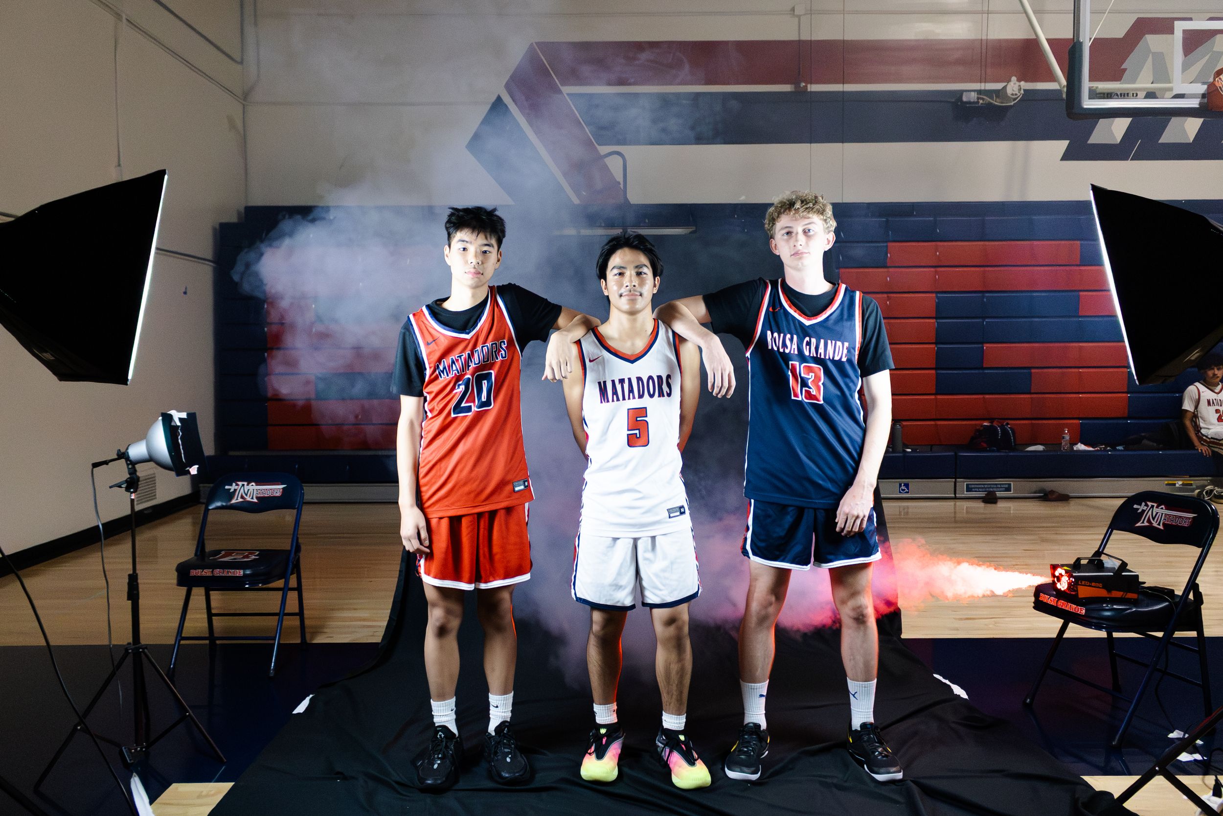 Three high school basketball players pose in team uniforms during a photo shoot with dramatic lighting and smoke effects.