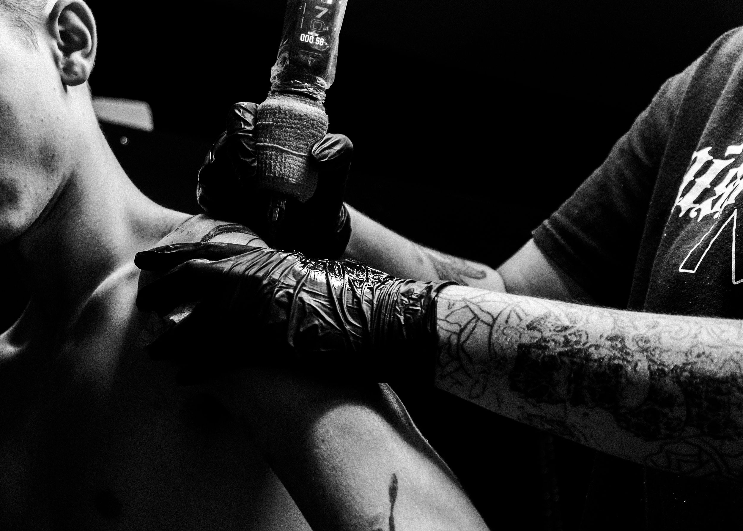 Black and white photo of a tattoo artist wearing gloves applying ink to a client's shoulder.