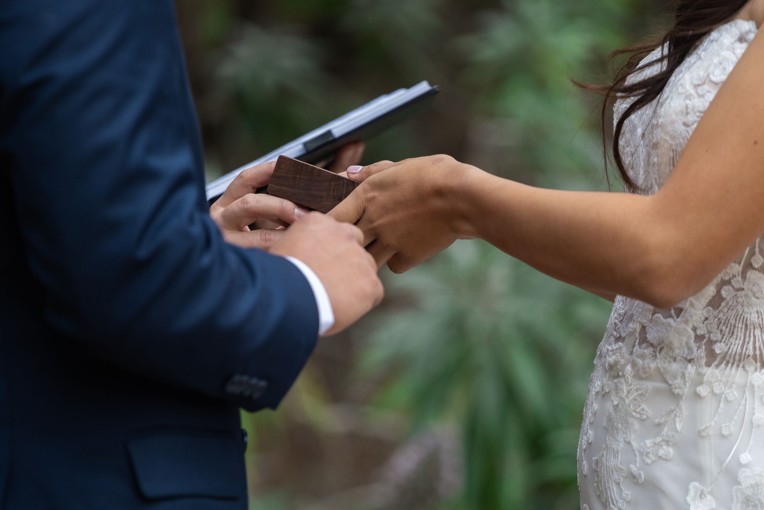 Bride and groom exchanging rings during outdoor wedding ceremony, close-up of hands holding ring box.