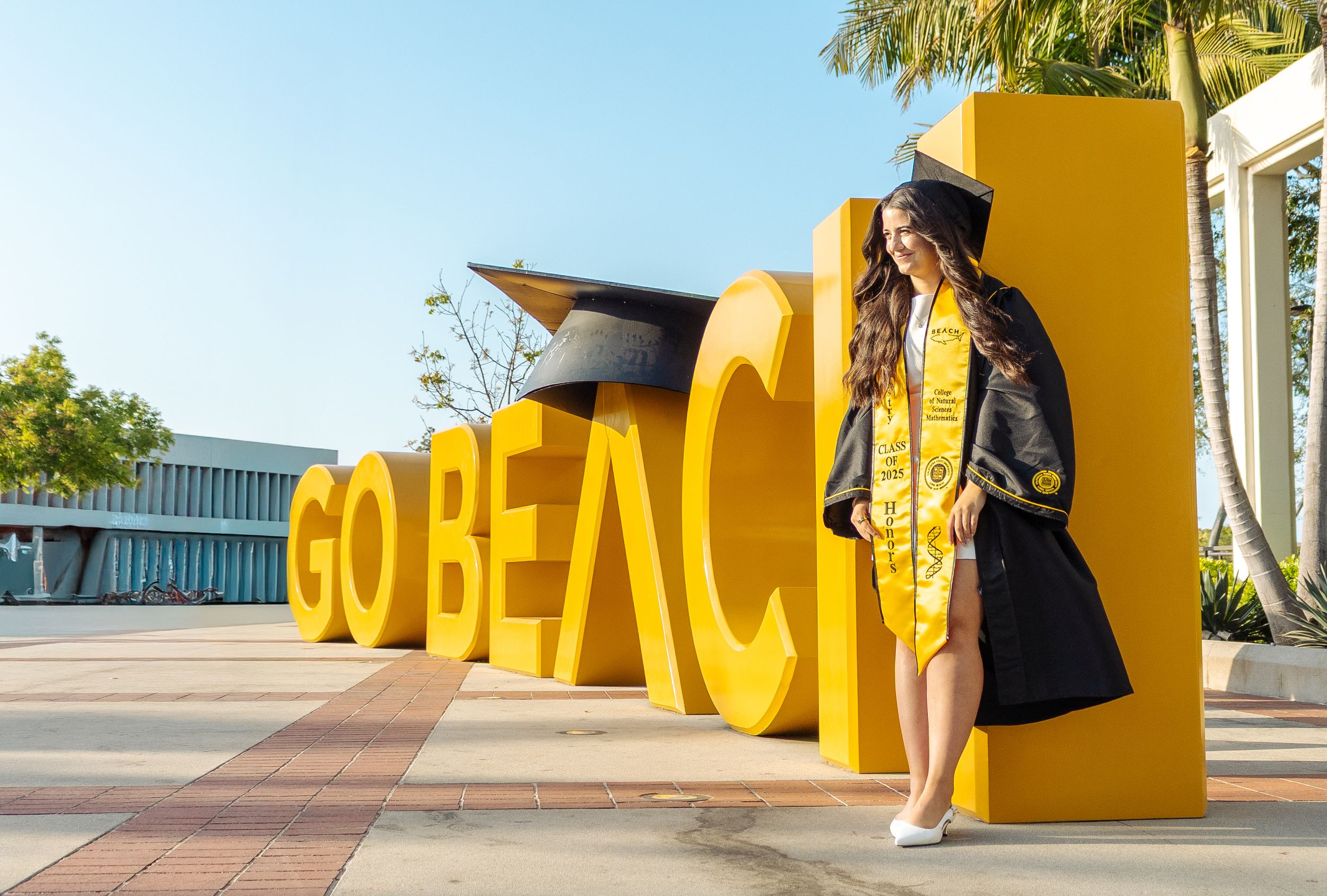 Graduate in cap and gown poses by yellow GO BEACH letters at Long Beach State University campus.