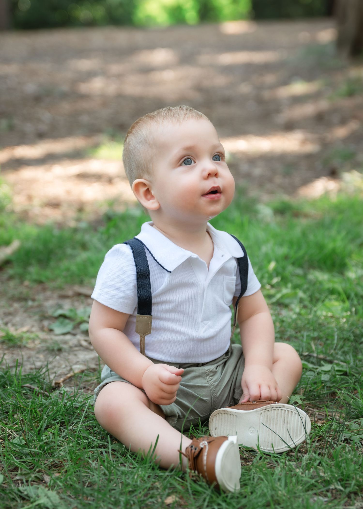 A cute baby boy in white polo shirt with suspenders and khaki shorts sits on grass in a sunny park.