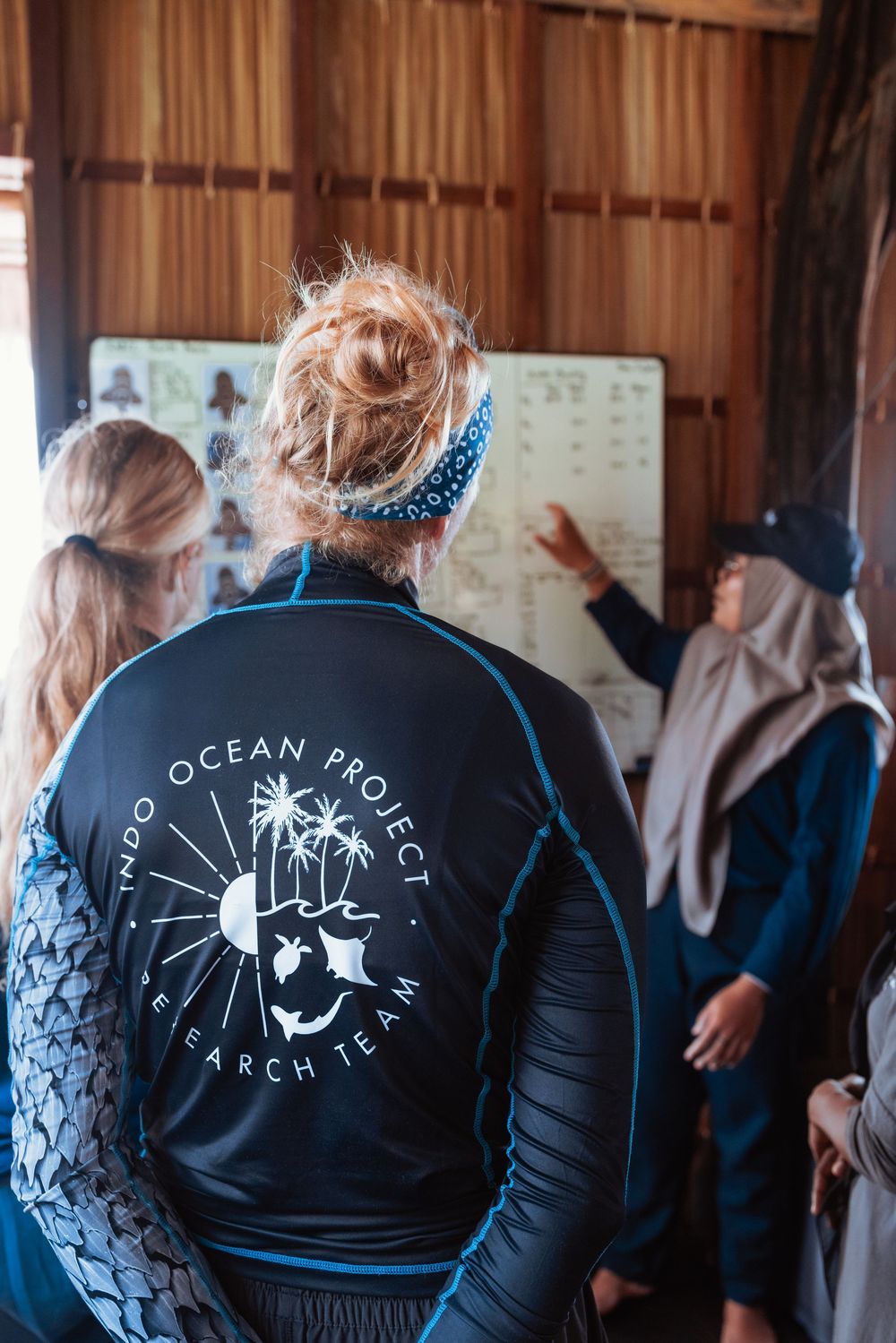 Researcher wearing Indo Ocean Project Search Team jacket points at marine life chart during team briefing.