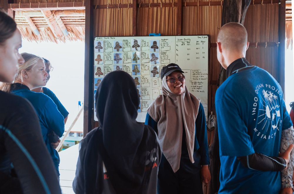 Group of people listening to a presenter in a tropical open-air building with a photo board display.