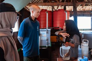 People conversing in a rustic open-air building with red water tanks, discussing water filtration equipment.
