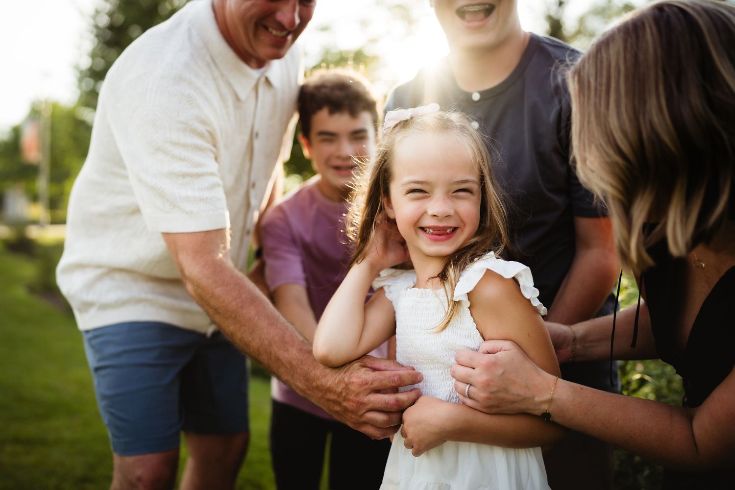 A joyful young girl in a white dress laughs surrounded by loving family members outdoors in a sunny green park.