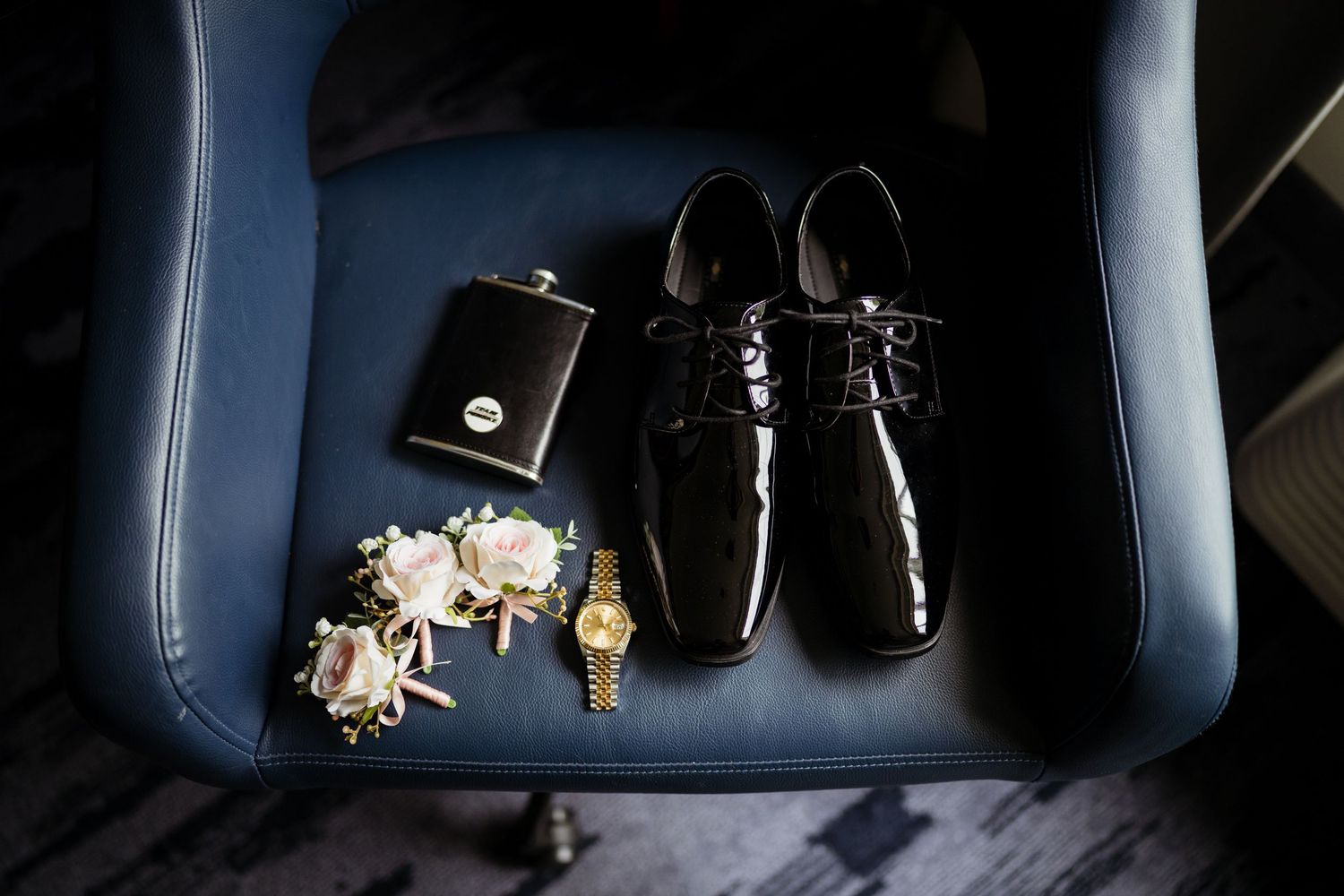 Black patent leather dress shoes, boutonniere, gold watch, and hip flask arranged on a blue leather chair for a wedding.