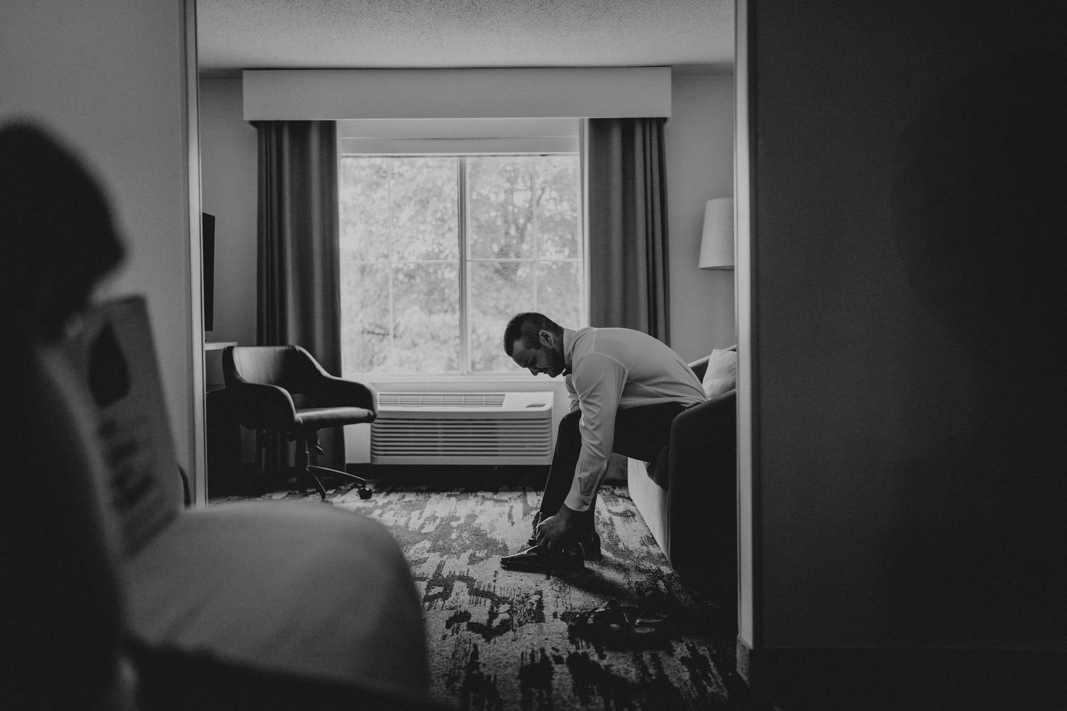 Black and white photo of a groom sitting on a hotel bed, putting on his shoes while getting ready.