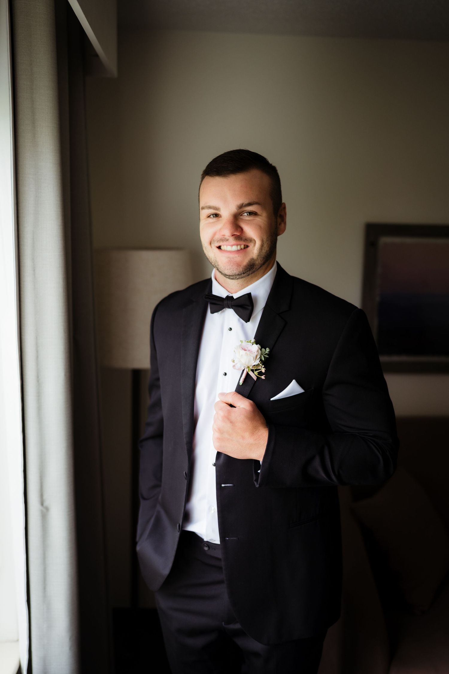 Smiling groom in black tuxedo with bow tie and boutonniere stands by window in hotel room.