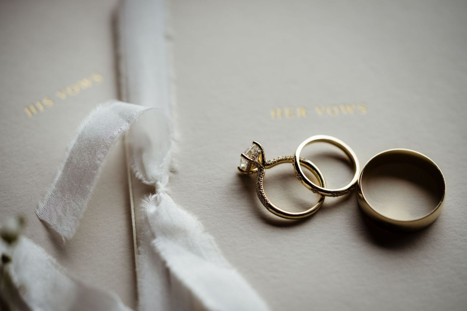 Gold wedding bands and diamond engagement ring resting on elegant vow books with white ribbon.
