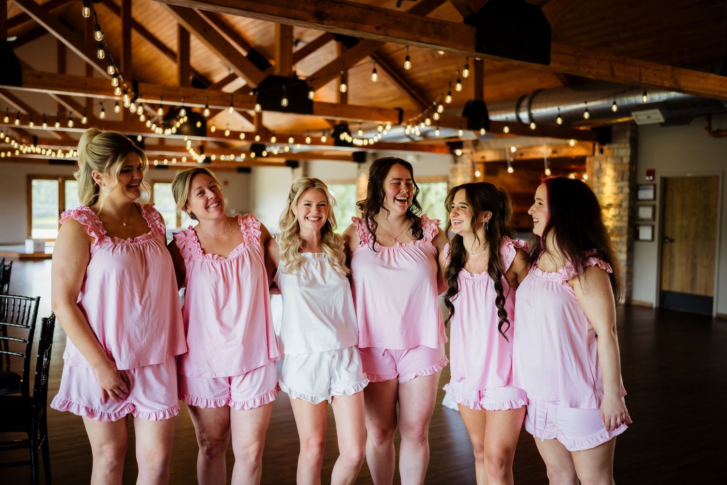 Six bridesmaids in matching pink pajama sets laughing together in a rustic barn venue with string lights.