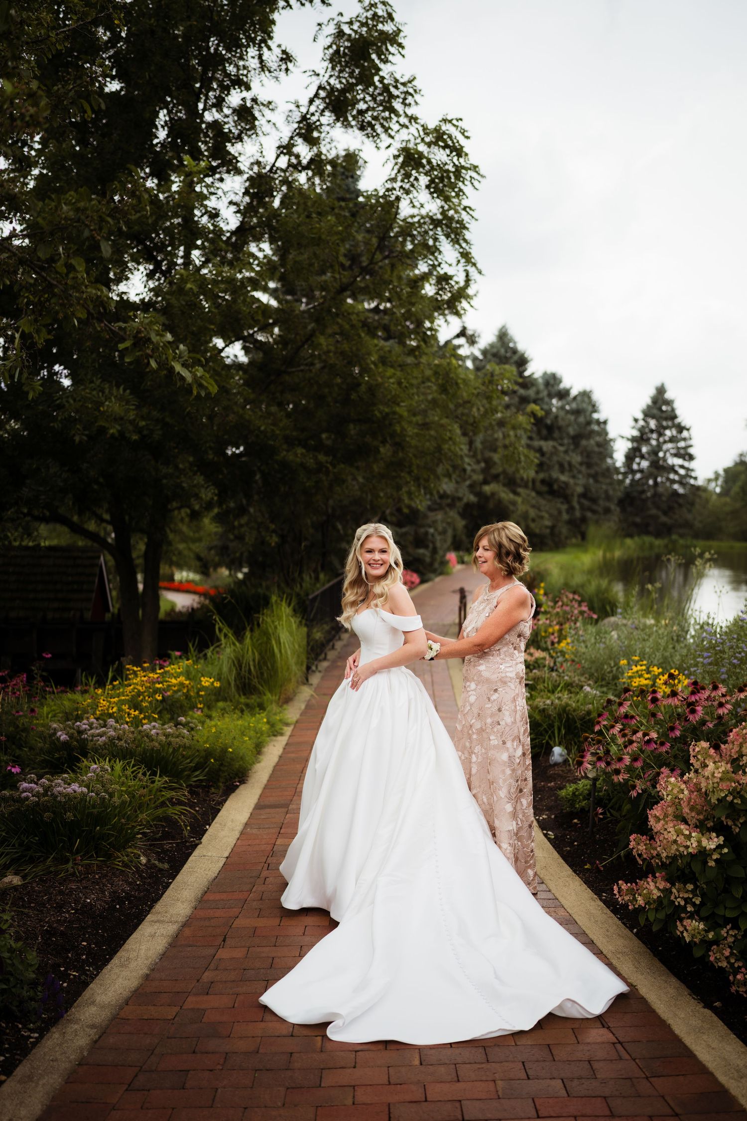 Bride in white off-shoulder ball gown with woman in floral dress on brick garden path surrounded by colorful flowers.
