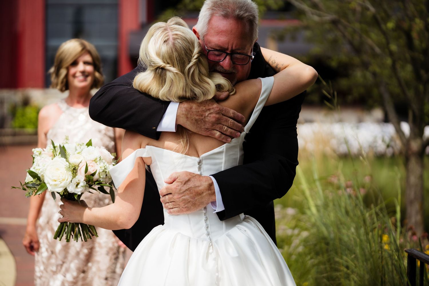 Emotional father-daughter wedding hug, bride in white gown holding bouquet, outdoor ceremony venue backdrop.