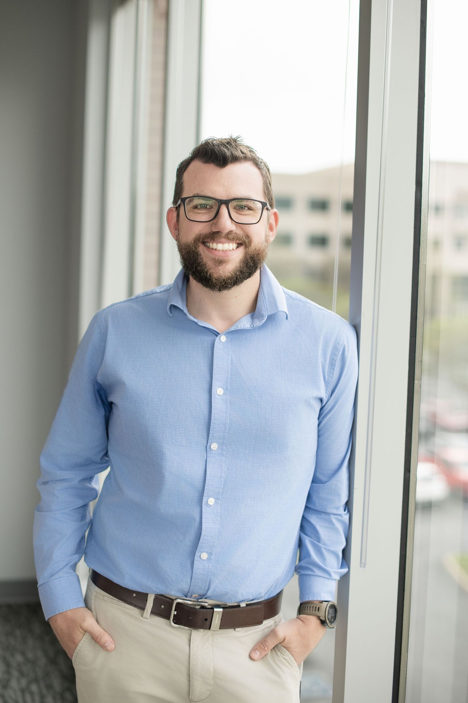 Headshot of smiling bearded man with glasses in blue shirt stands by office window in professional business portrait.