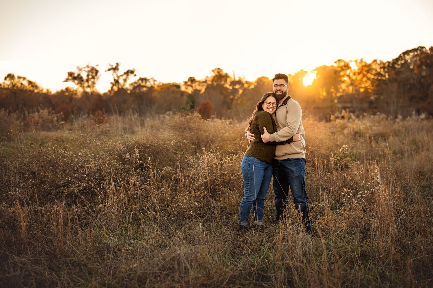 Couple embracing in golden field at sunset, surrounded by tall grass and trees in warm autumn light.