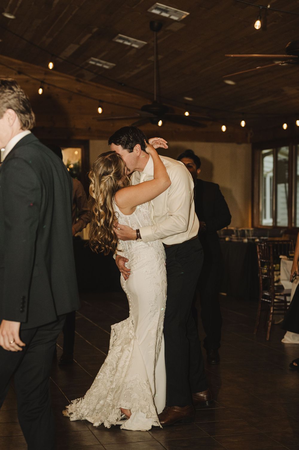 Bride and groom sharing first dance kiss in warmly lit rustic wedding reception venue.