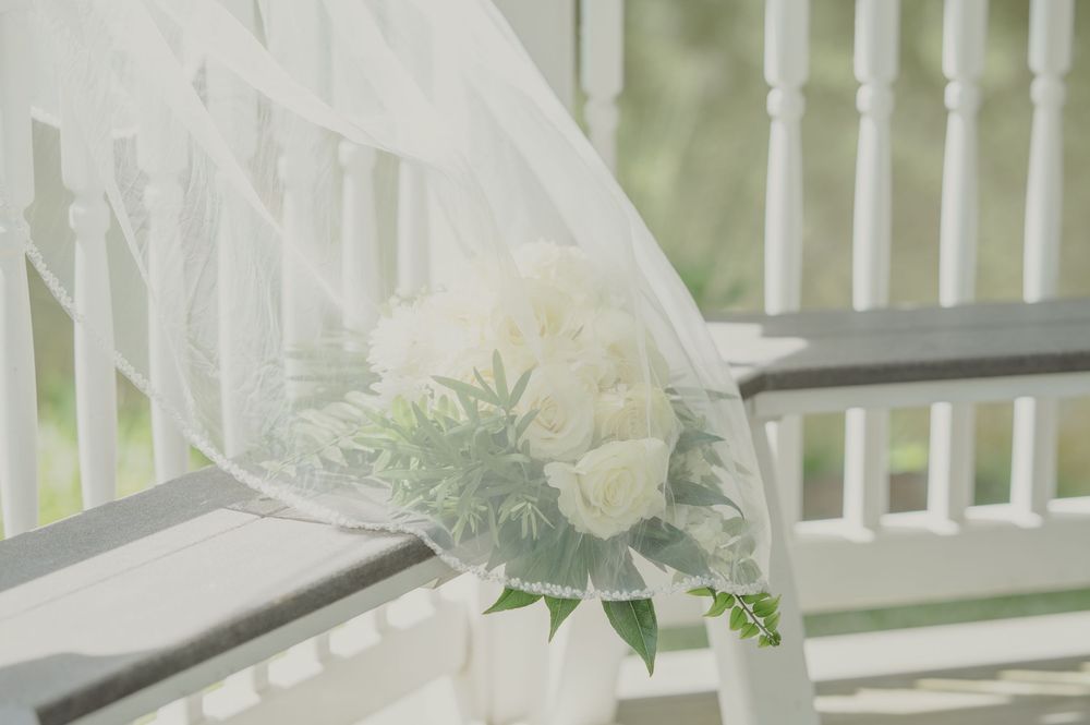 White bridal bouquet with roses and greenery draped with sheer veil on white staircase railing.